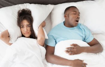 Loudly snoring man sleeping next to an annoyed woman covering her ears wih a pillow.